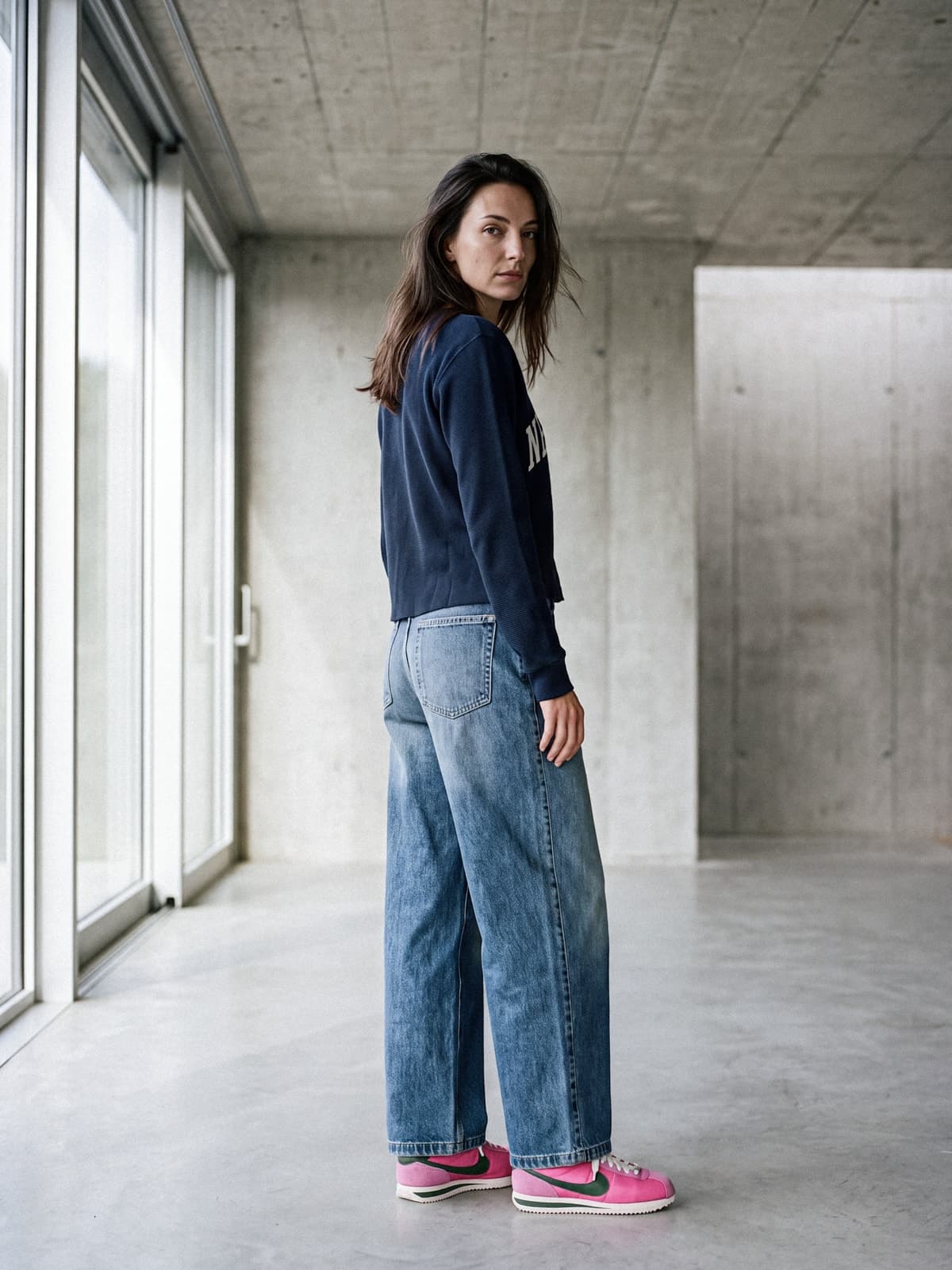 La mannequin de dos, tournée vers l'objectif, en sweat bleu marine, jean ample et baskets roses, dans un intérieur de béton bordé de baies vitrées.