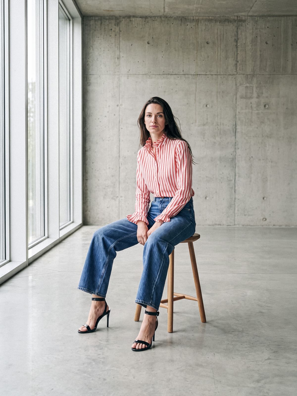 La mannequin assise sur un tabouret en bois, en chemise à fines rayures rouges et blanches, jean ample et sandales noires à talons, devant une baie vitrée.