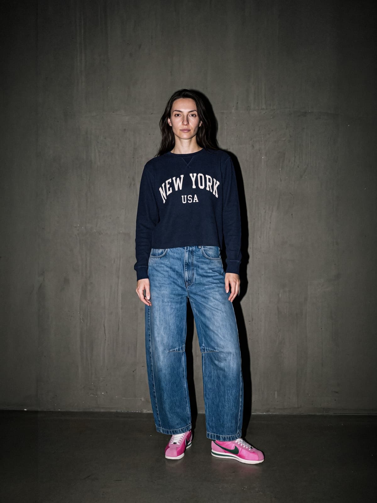La mannequin en sweat bleu marine floqué NEW YORK USA, jean ample et baskets roses, devant un mur de béton sombre.